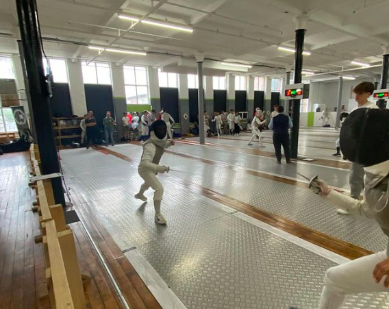 Two girls fencing on electric piste at a busy fencing competition.